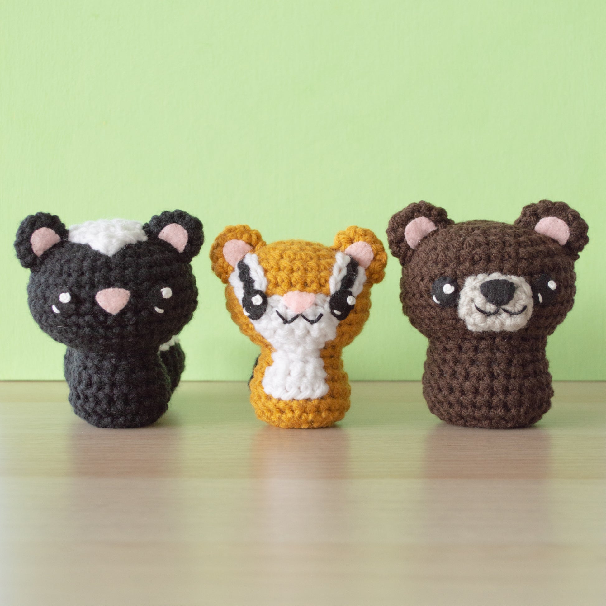 Three delightful crochet amigurumi figures are displayed on a wooden surface against a light green background. From left to right: a charming skunk crochet creation, a playful tan and white tiger, and an adorable bear crochet masterpiece.