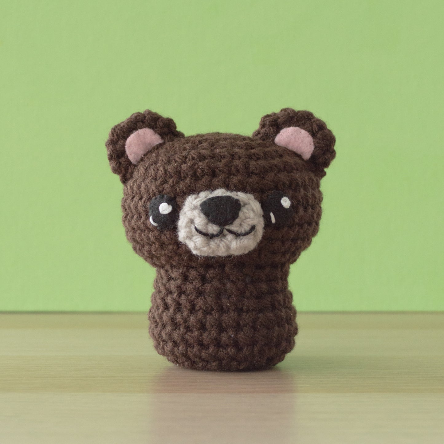 A cute, brown bear crochet with pink inner ears and a smiling face sits on a wooden surface. Expertly crafted using a crochet amigurumi pattern, it pops against the light green wall in the background.