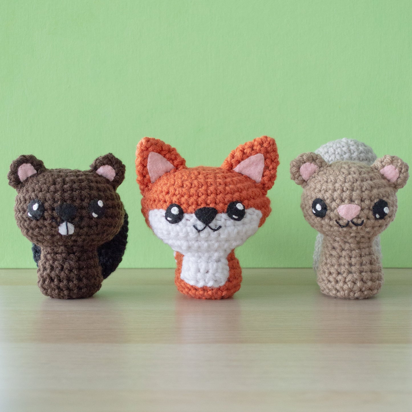 Three charming backyard buddies, crafted using a crochet amigurumi pattern, stand in a row on a light wood surface against a green background. From left to right: a brown beaver, an orange and white fox, and a light brown squirrel with a gray tail bring wild whimsy to any setting.