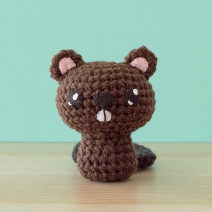 A cute crochet amigurumi pattern of a stuffed animal sits on a wooden surface against a light green background. The toy is brown with pink inner ears, white eyes, and a small pink mouth, ready to join your collection of backyard buddies.