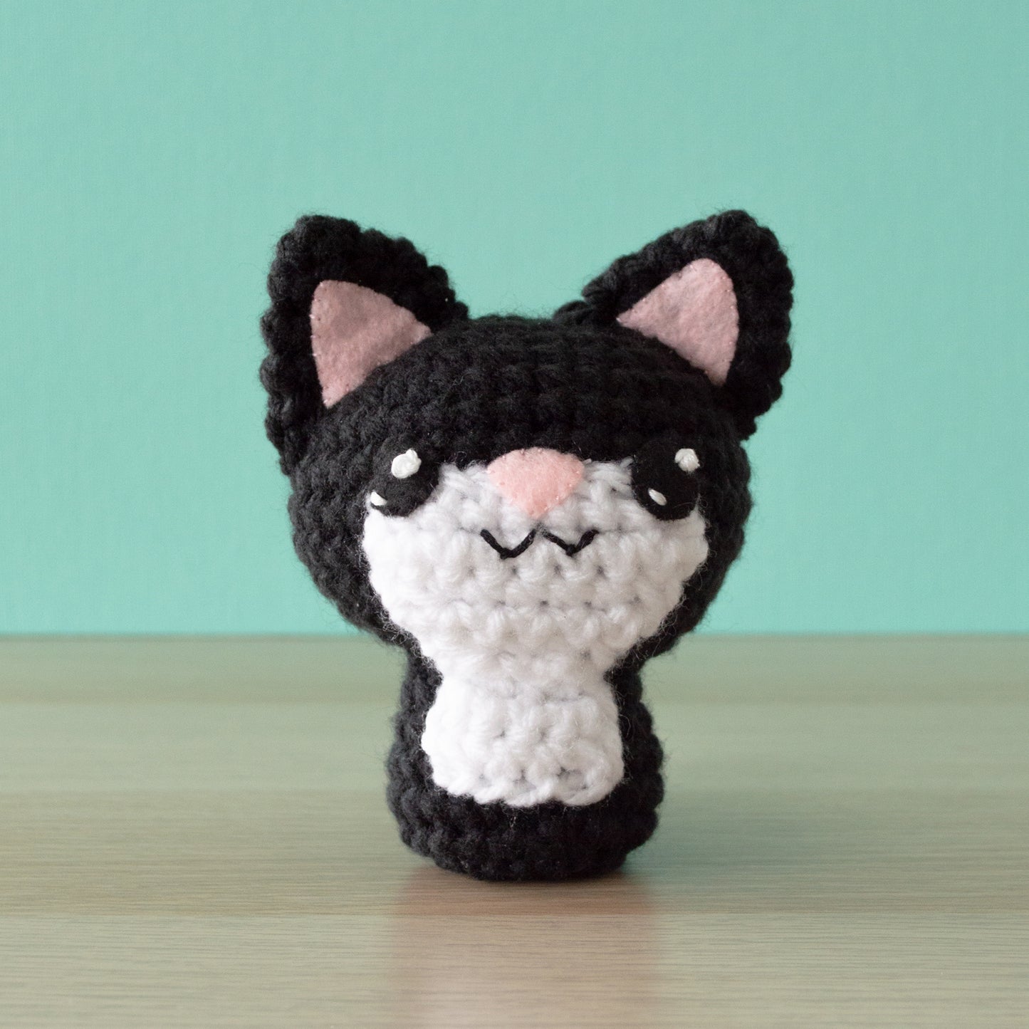 A small crochet amigurumi black tuxedo cat with a white chin and belly, a pink nose and big black eyes, sitting on a wooden surface with a blue background.