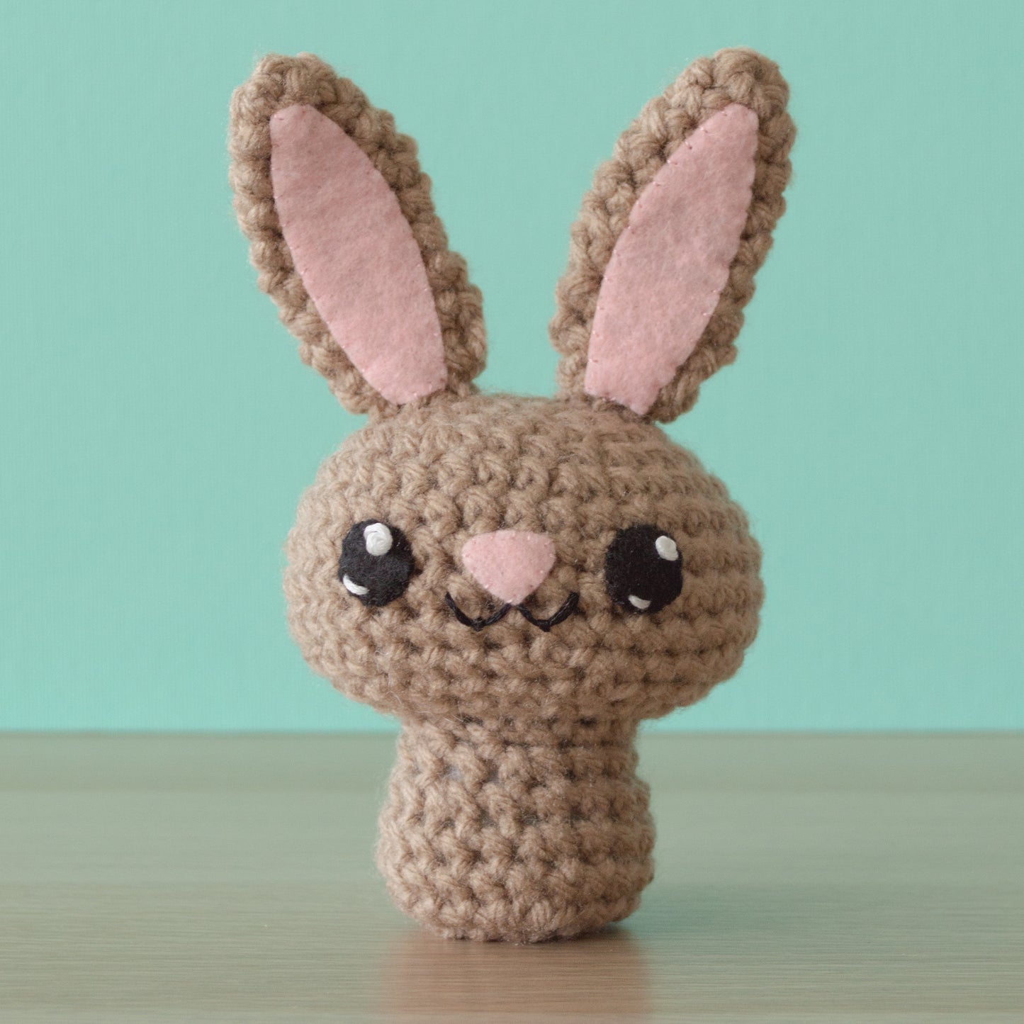 A charming crocheted bunny, part of a delightful crochet menagerie, showcases large pink ears and a cute face with bright eyes and a small nose, all set against a turquoise background.