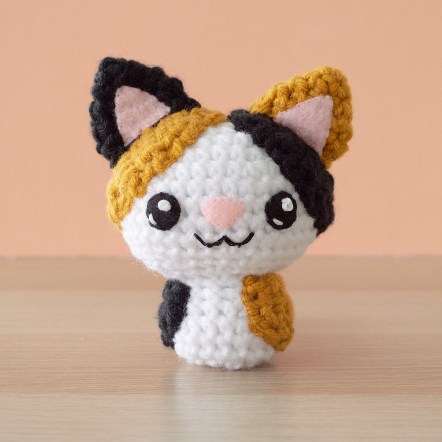 A small crochet amigurumi white calico cat with black and gold spots, a pink nose and big black eyes, sitting on a wooden surface with an orange background.