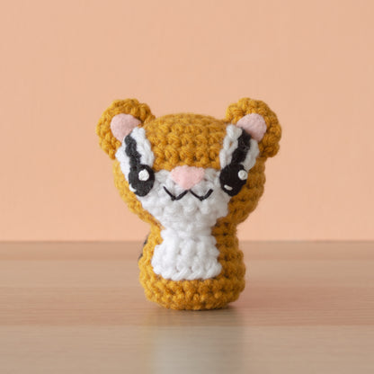 A charming crochet amigurumi pattern, featuring a small toy resembling a squirrel, sits on a wooden surface. The handcrafted piece is orange with white and black facial details and a white chest. The pale peach background perfectly complements its vibrant hues.