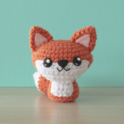 A cute crocheted fox amigurumi, crafted from orange and white yarn, sits on a wooden surface against a teal background. With large black eyes, a small nose, and a cheerful expression, this little creature embodies wild whimsy in its delightful design.