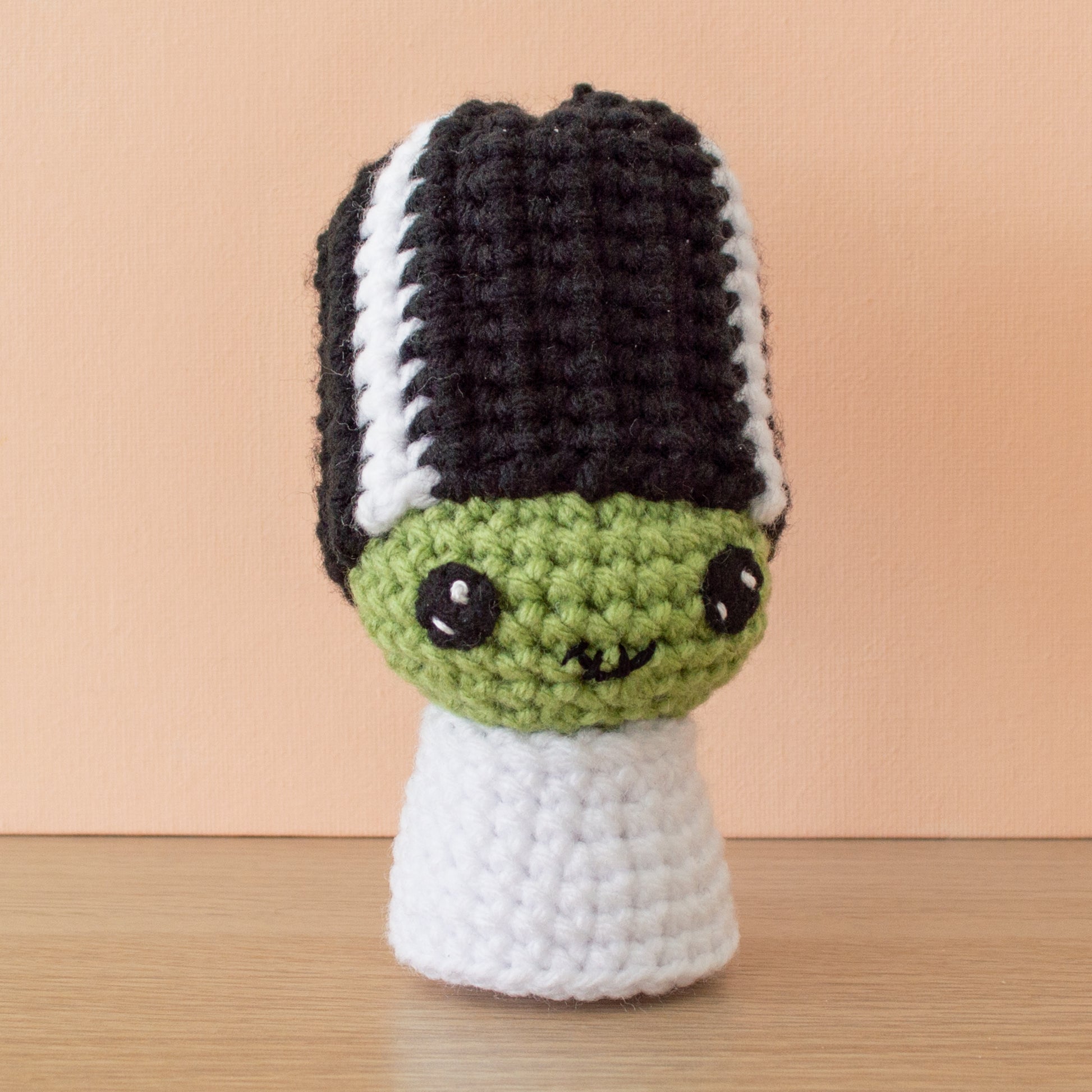 A small green crochet amigurumi Bride of Frankenstein with tall black-and-white striped hair and a stitched smile displayed on a wooden surface against a peach-colored background.