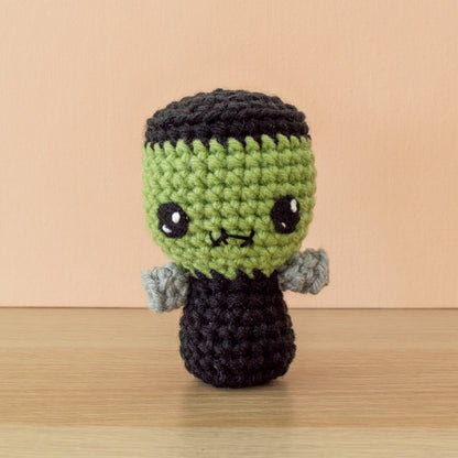 A small thin crocheted green Frankenstein monster with black hair dressed in a simple black suit with silver bolts on each side of his neck and a downturned stitched frown.