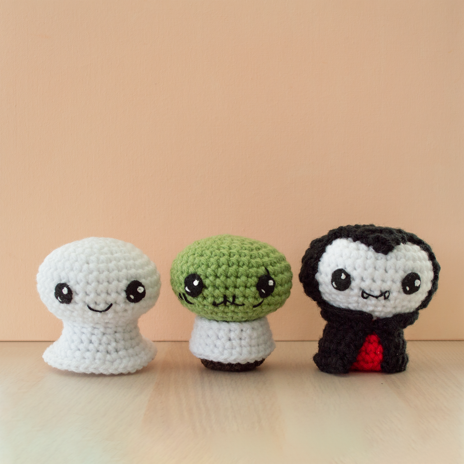 Three small crochet amigurumi characters lined up on a wooden surface against a peach-colored background. From left to right: a white ghost with large black eyes and a small smile, a green zombie with black eyes, stitched mouth details, and a round head, and a vampire with pale white skin, black hair, a red shirt, and a black cape with a small fang-toothed smile.