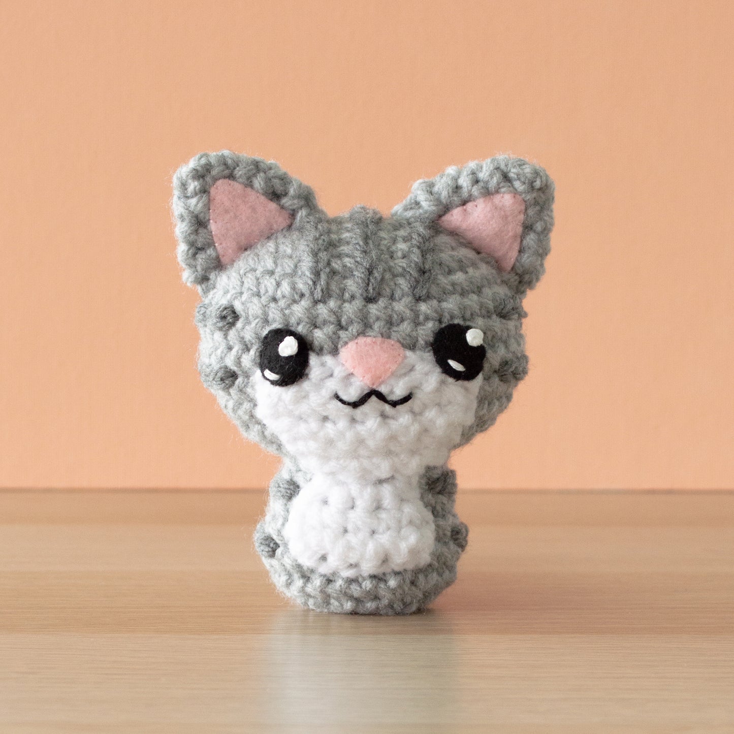 A small crochet amigurumi gray tabby cat with drank gray stripes, a white chin and belly, a pink nose and big black eyes, sitting on a wooden surface with an orange background.