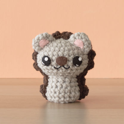 A small, crocheted hedgehog with big black eyes, a stitched smile, and light brown and dark brown detailing sits on a wooden surface against a peach background. This adorable creature is part of a charming crochet menagerie, perfect for those seeking backyard-inspired whimsy in their decor.