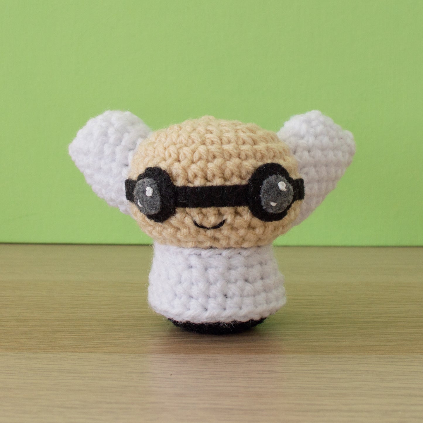 A small crochet amigurumi scientist with beige skin, black round glasses, white hair, and a white lab coat displayed on a wooden surface against a peach-colored background.