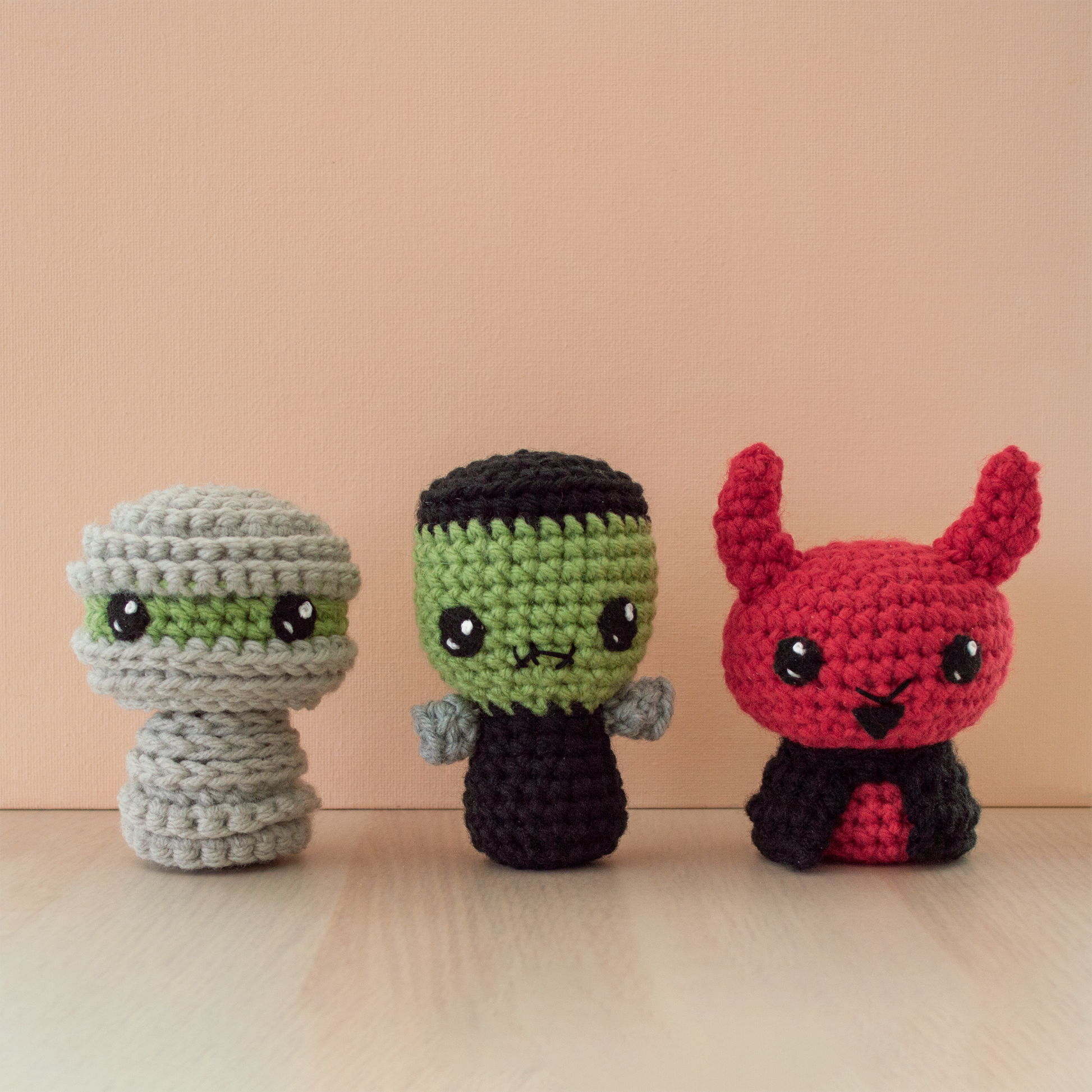 Three small crochet amigurumi characters displayed on a wooden surface against a peach-colored background. From left to right: a gray mummy wrapped in textured layers with large black eyes, a green Frankenstein monster with black hair, a stitched mouth, and gray bolts, and a red devil with upright horns, black eyes, a small smile, goatee, and a black cape.