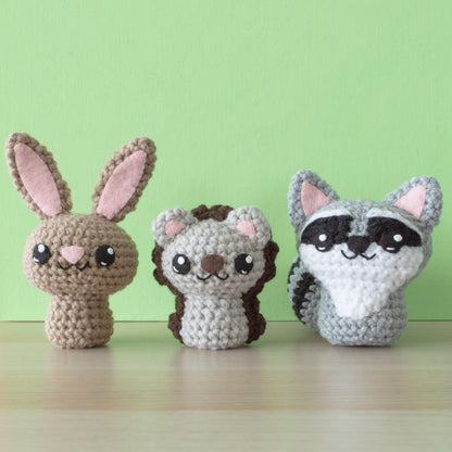 Three cute crocheted animal figures, like a backyard-inspired crochet menagerie, rest on a wooden surface against a light green background: a brown bunny with pink ears, a hedgehog with a brown back, and a gray raccoon with a black mask around its eyes.