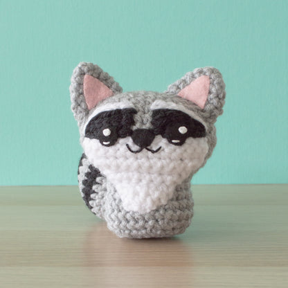 A backyard-inspired crocheted raccoon plushie, with pink ears and a smiling face, sits on a wooden surface against a mint green background.