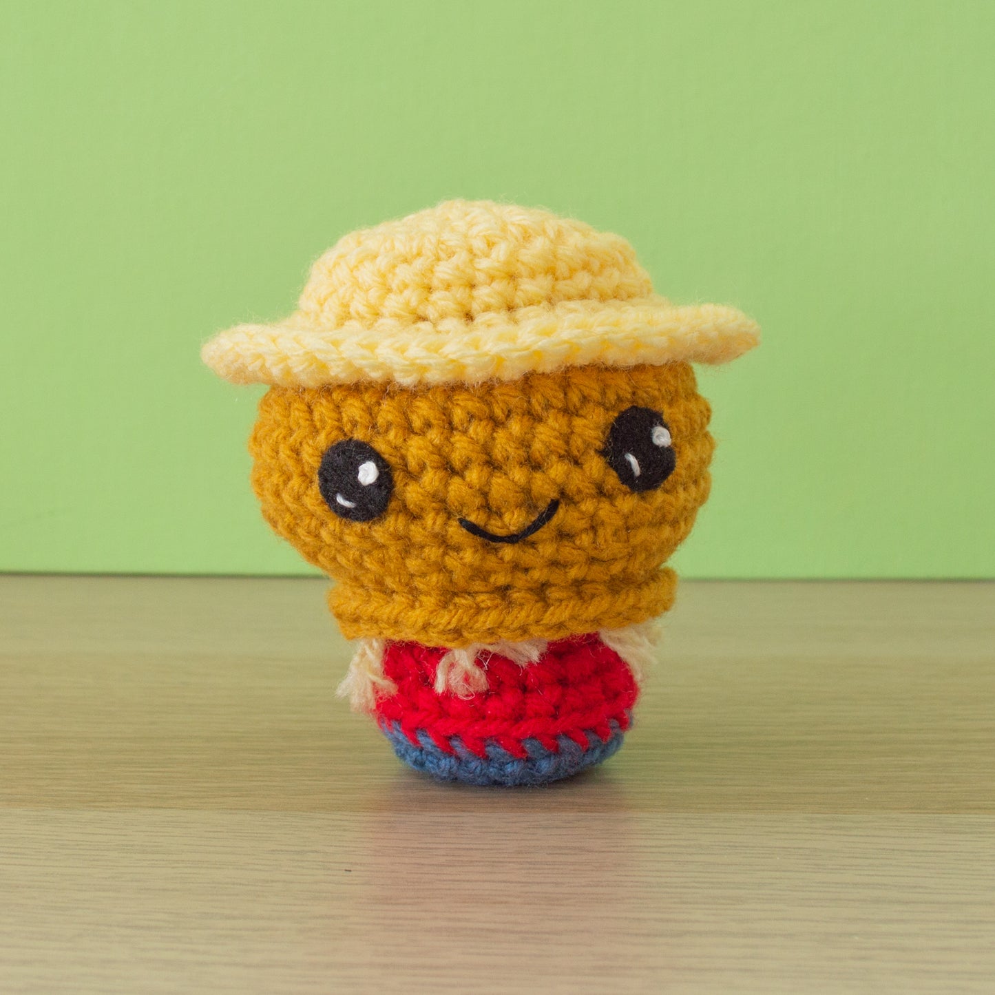 A small crocheted scarecrow with a gold head with straw at the neck, dressed in a red shirt and blue pants wearing a wide brimmed straw-yellow hat with large black eyes and a small smile.