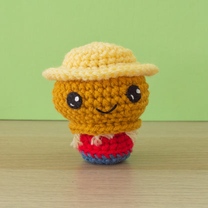 A small crocheted scarecrow with a gold head with straw at the neck, dressed in a red shirt and blue pants wearing a wide brimmed straw-yellow hat with large black eyes and a small smile.