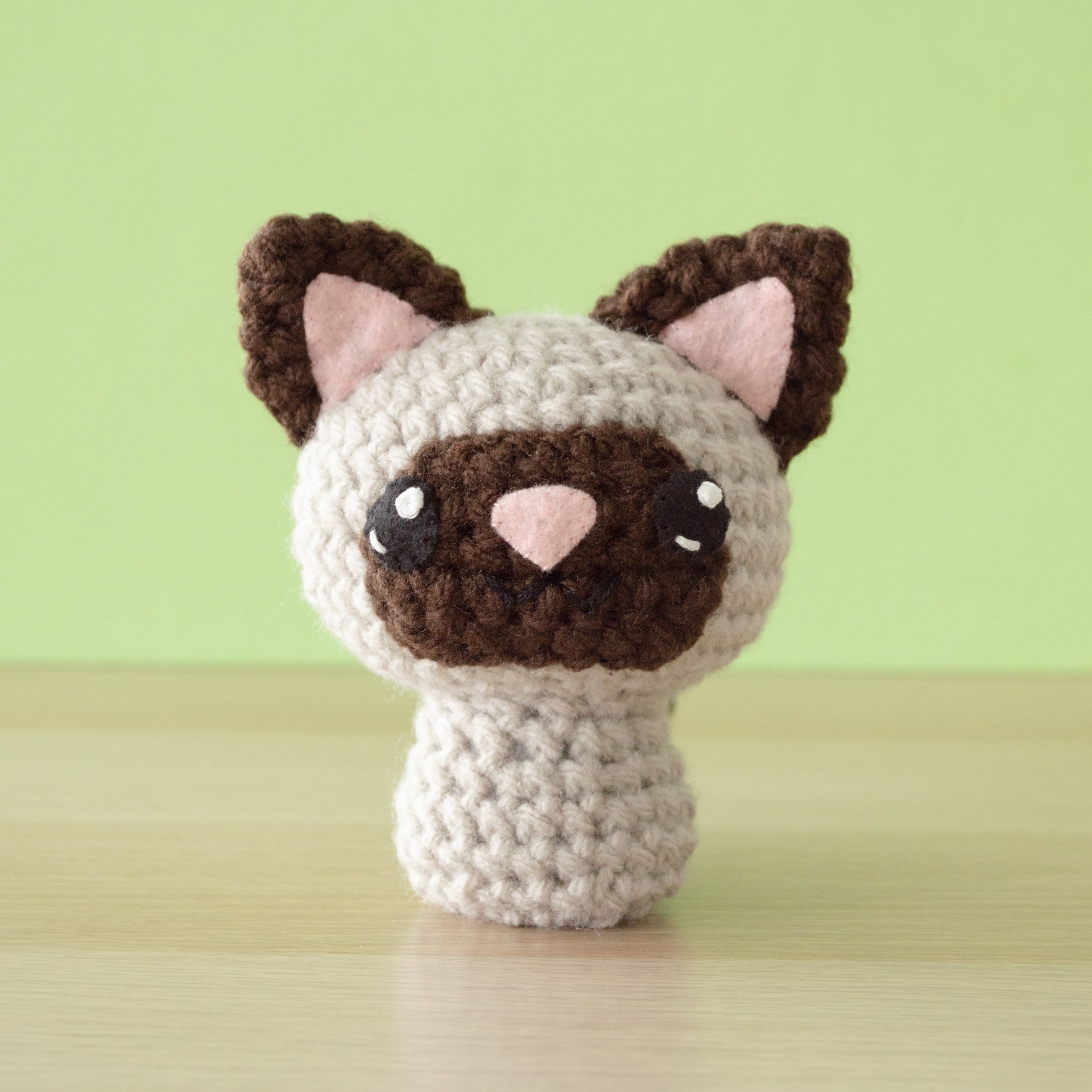 A small crochet amigurumi cream-colored siamese cat with brown points, a pink nose and big black eyes, sitting on a wooden surface with a green background.