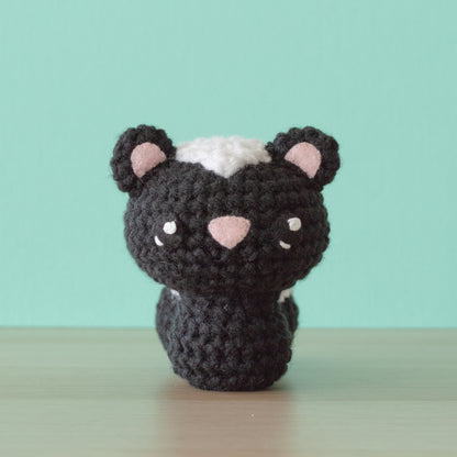 A skunk crochet amigurumi with a black body, white stripe on its head, and pink accents on its ears sits on a wooden surface against a turquoise background.