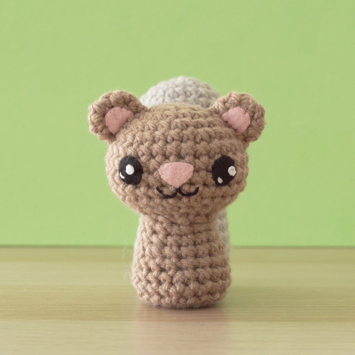 A crocheted brown and beige bear-like creature with big eyes and pink ears sits on a wooden surface against a green background, bringing a touch of wild whimsy to your backyard buddies collection.