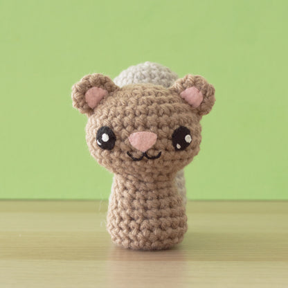 A crocheted brown and beige bear-like creature with big eyes and pink ears sits on a wooden surface against a green background, bringing a touch of wild whimsy to your backyard buddies collection.