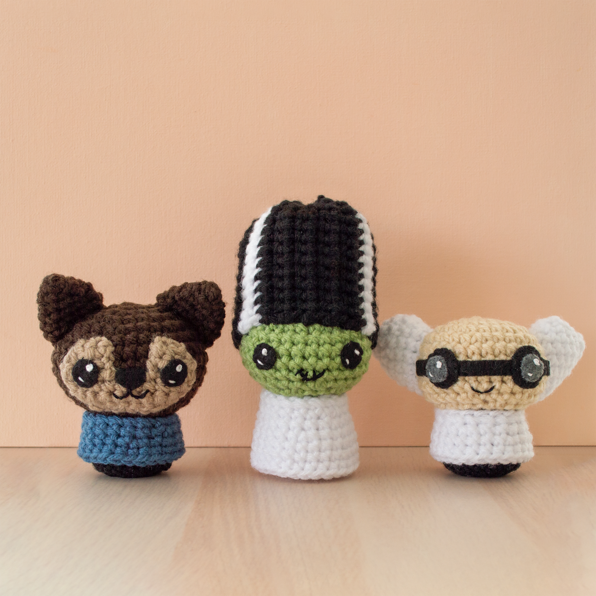 Three small crochet amigurumi characters displayed on a wooden surface against a peach-colored background. From left to right: a brown werewolf with pointy ears, beige face markings, a blue shirt, and a small smile; a green Bride of Frankenstein with tall black-and-white striped hair and a stitched smile; and a scientist with beige skin, black round goggles, white hair, and a white lab coat.