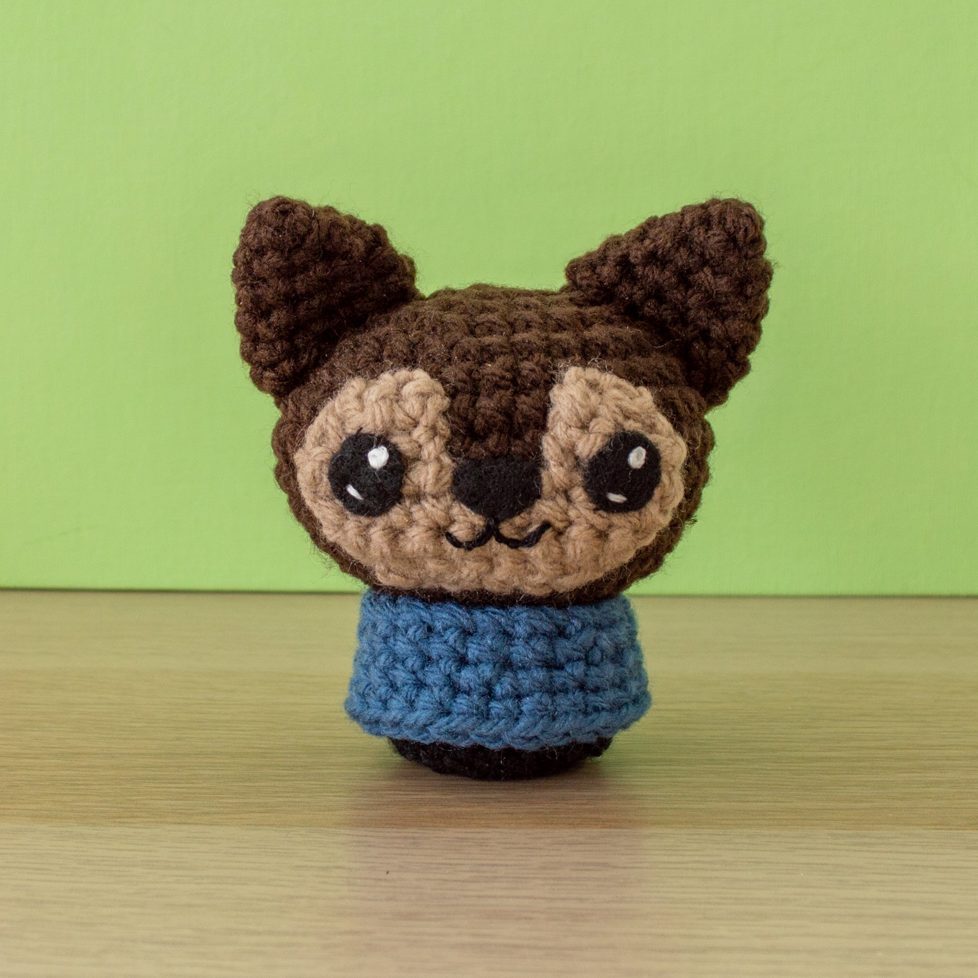 A small brown crochet amigurumi werewolf with pointy ears, beige face markings, a blue shirt, and a small smile displayed on a wooden surface against a peach-colored background.