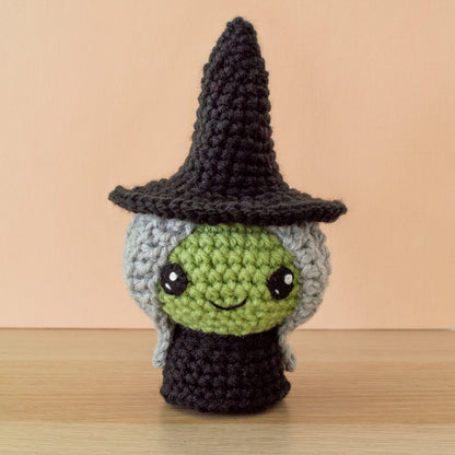 A small crocheted green witch dressed in a simple black dress, tall pointy black hat with long crocheted gray hair with large black eyes and a small smile.