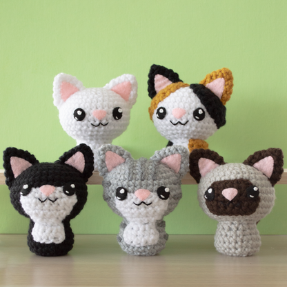 A collection of small crochet amigurumi cats sitting on a wooden surface with a green background. In the back row sitting on a wooden board from left to right is a white kitty and a calico kitty with gold and black spots. In the front from left to right is a black tuxedo kitty with a white face and belly; a gray tabby kitty with dark gray stripes and a white face and belly; a cream colored siamese cat with brown ears and face.